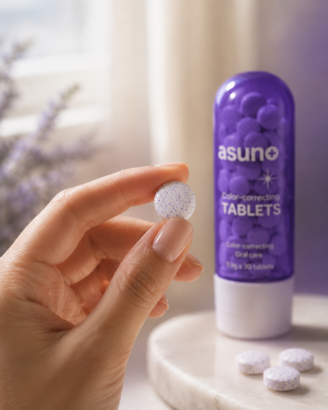 Hand holding a white, lavender-speckled Asuno Color-Correcting Tablet beside the violet-tinted bottle and glass of water